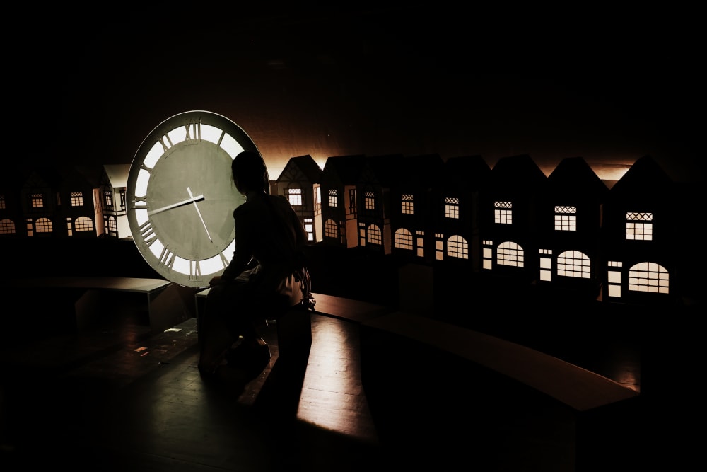 Image shows a silhouette of a person sitting in front of a large clock with small lit up houses in the background 