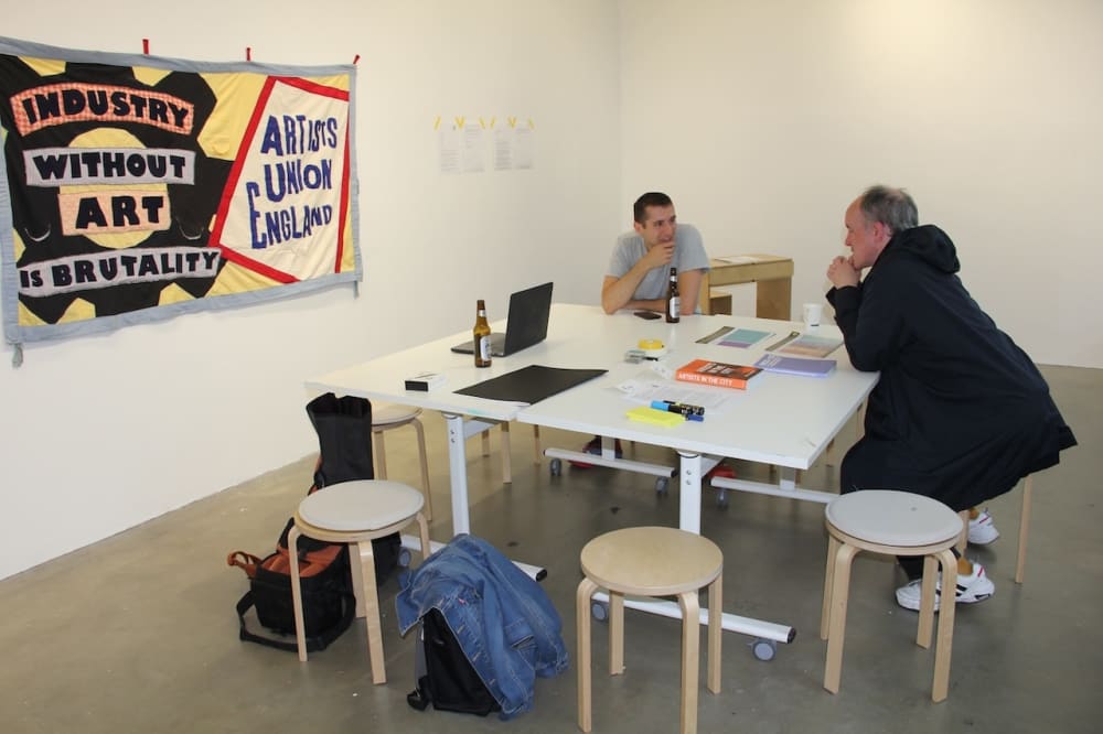 Image shows 2 people sitting on stools round a table in a large white room, seemingly in conversation. In the background, a banner can be seen hanging on the wall which reads “Industry without art is brutality. Artists’ Union England”