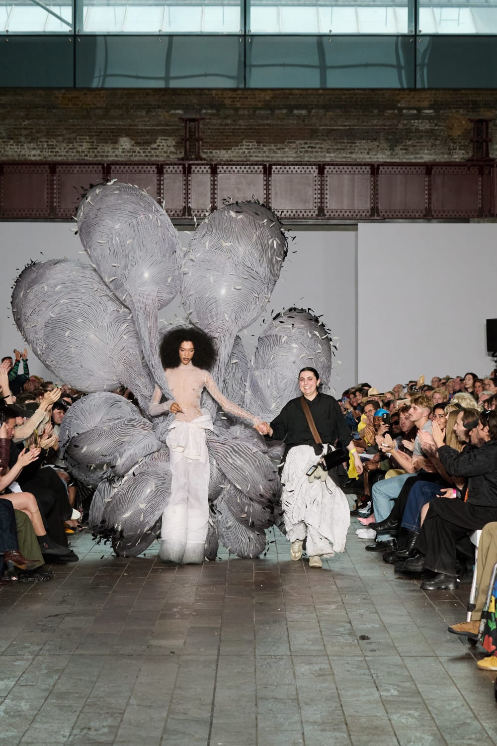 Model and designer walking down a catwalk with people sat on each side.