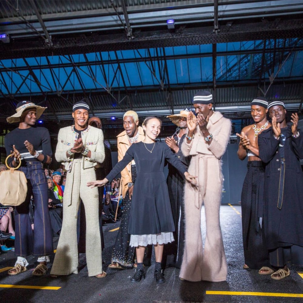 An image of multiple people on a runway. Some are clapping, the mood is celebratory