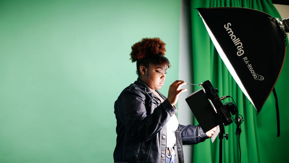 A person adjusts a lighting setup in front of a green screen, illuminated by a large softbox in a studio setting.
