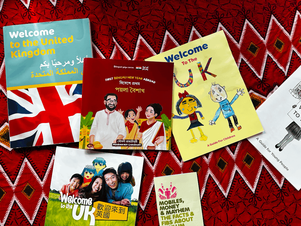 A collection of multilingual welcome guides and children's resources about living in the UK displayed on a red patterned fabric. At the center is a bilingual Bengali-English children's book, First Bengali New Year Abroad by Moubani Roy Choudhury, surrounded by guides in Arabic, Chinese, and English.