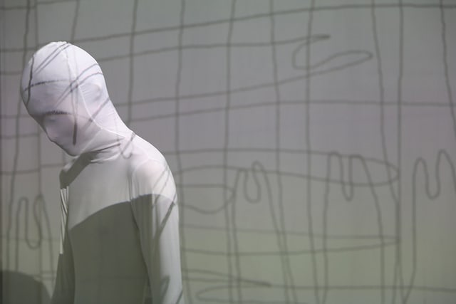 Figure all in white with their face covered and abstract shadows projected onto them.