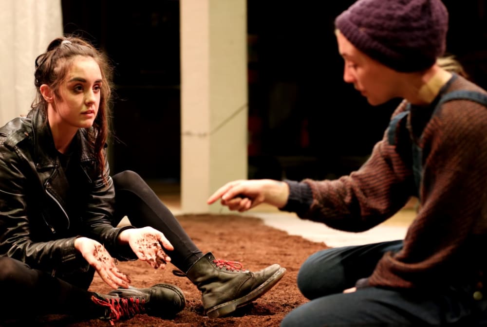 Two people on a stage, sitting in dirt.