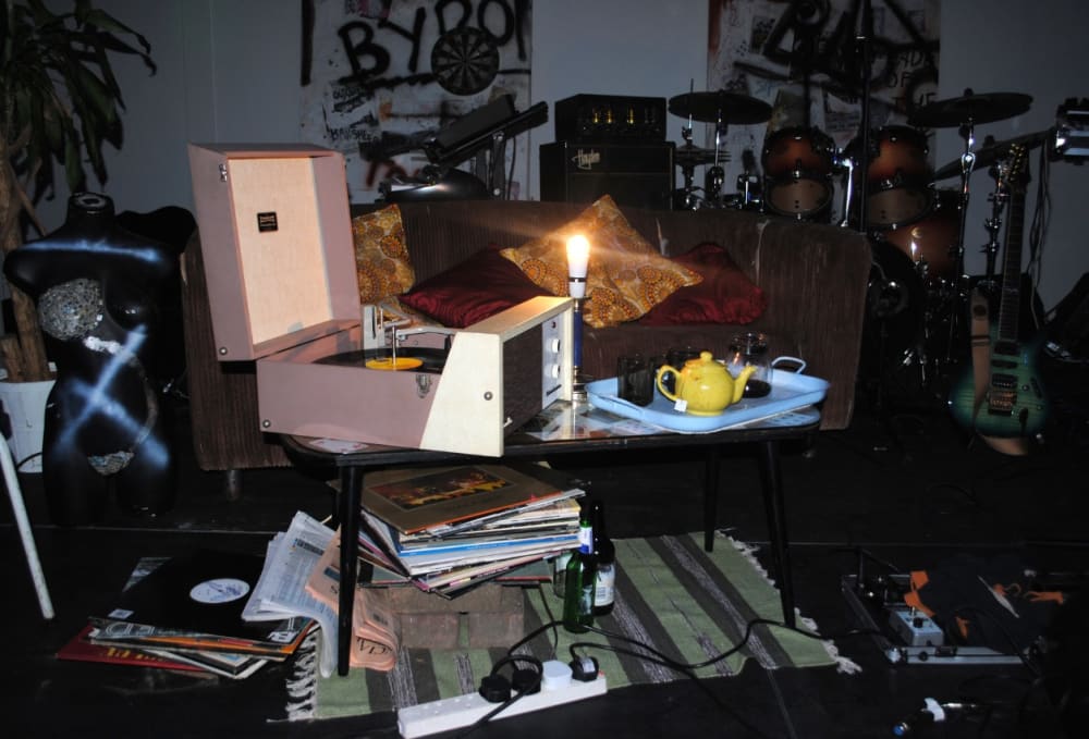A set featuring a pink record player on a coffee table surrounded by a teapot, a drum kit, records and beer bottles.