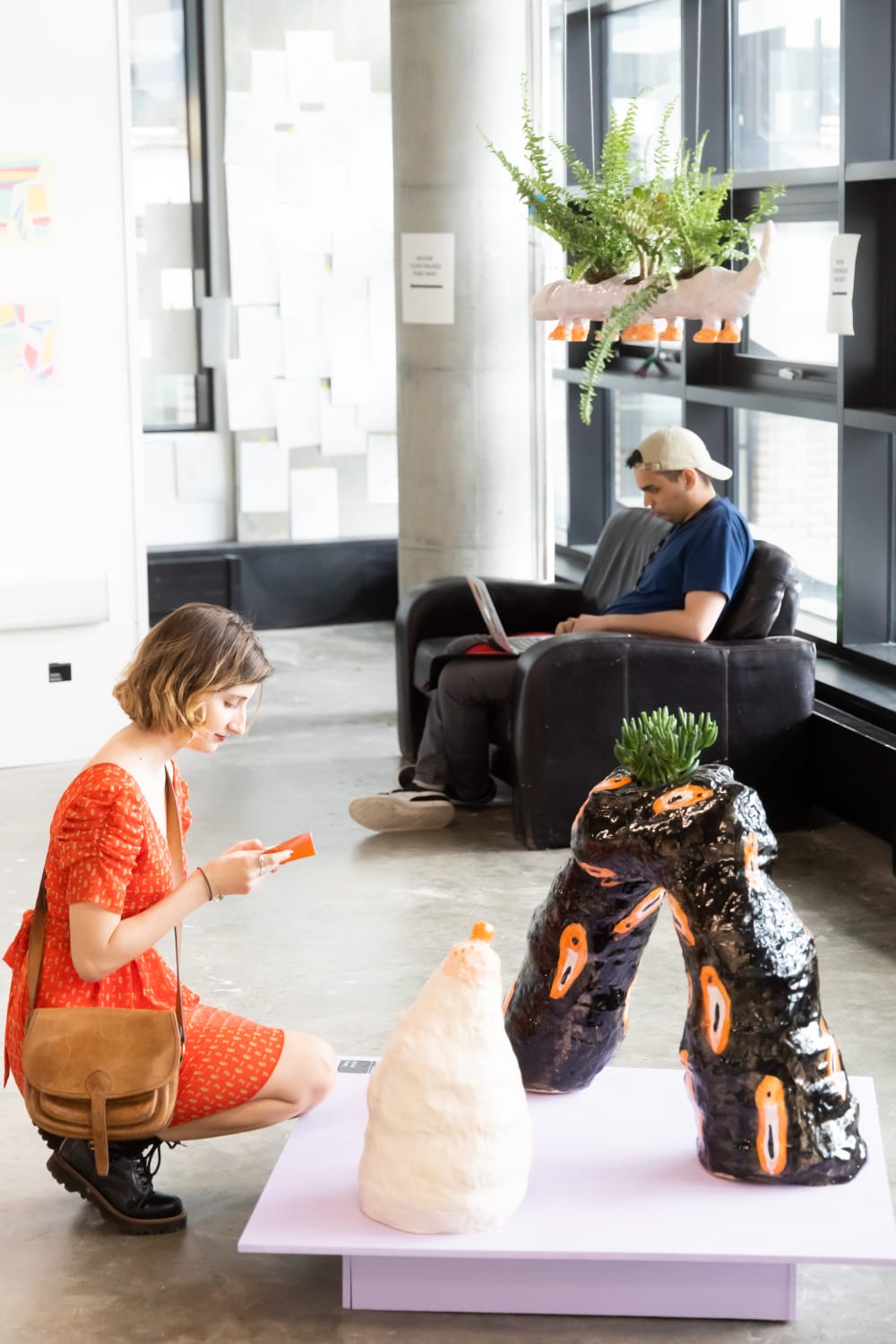A woman in an orange dress kneels down in from of ceramic sculptures on a lilac plinth. In the background, a man is sitting on a black sofa on his laptop. There is a ceramic sculpture hanging from the ceiling which is planted with ferns.