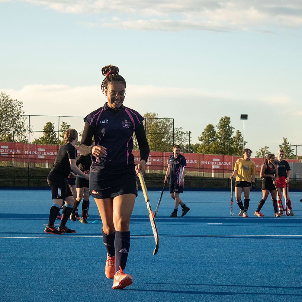 A photograph of a woman playing Hockey, she is wearing a UAL Hockey T-Shirt and is holding a hockey stick. Behind her are other members of her team. She is smiling and looking down at the pitch.