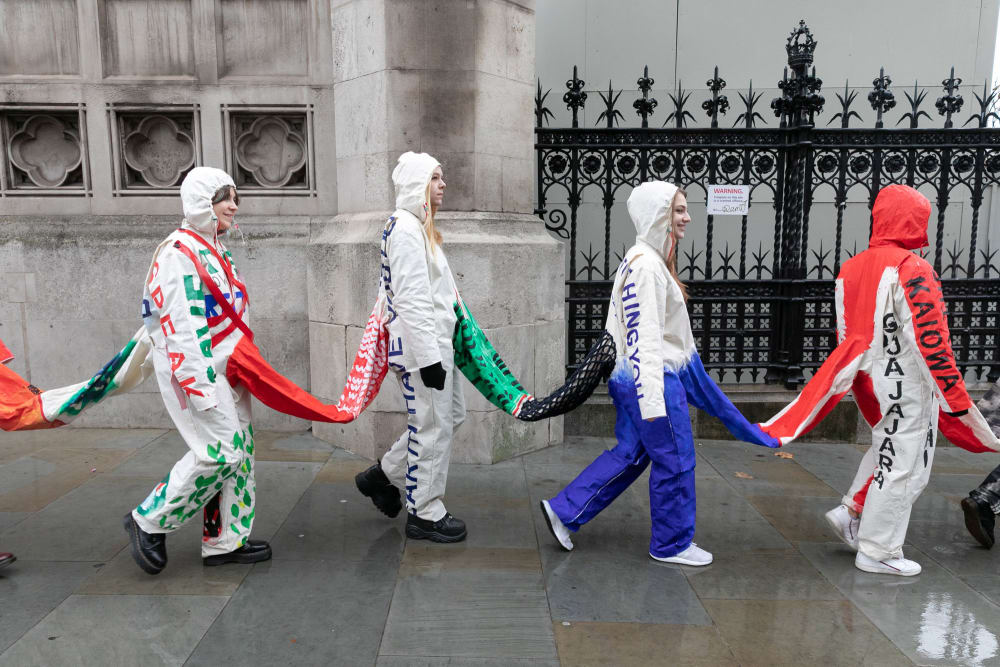 Four people, part of a larger chain, wearing joined suits walk on a city street past an old wall and ornate gate.