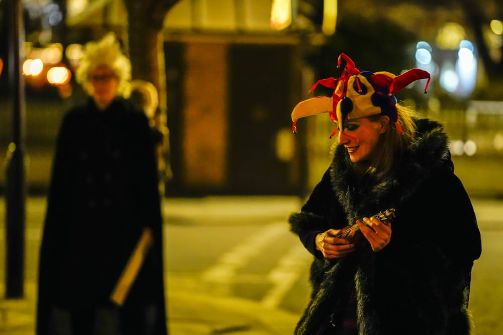 Two people standing in the street at night. The one to the left is blurry, and the woman to the right is wearing a jesters hat