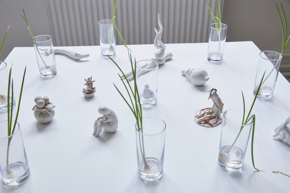 An installation piece, a series of ceramic models are displayed on a white tablecloth. Several glass vases hold grass. 