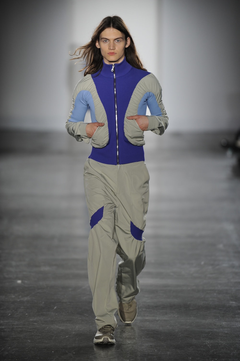 Man on runway wearing indigo, grey and pale blue silk fit short zip up bomber style jacket with ruched detailing and cuffed neck and sleeve, hand in pockets paired with grey straight pants with blue side upper calve detail and trainers
