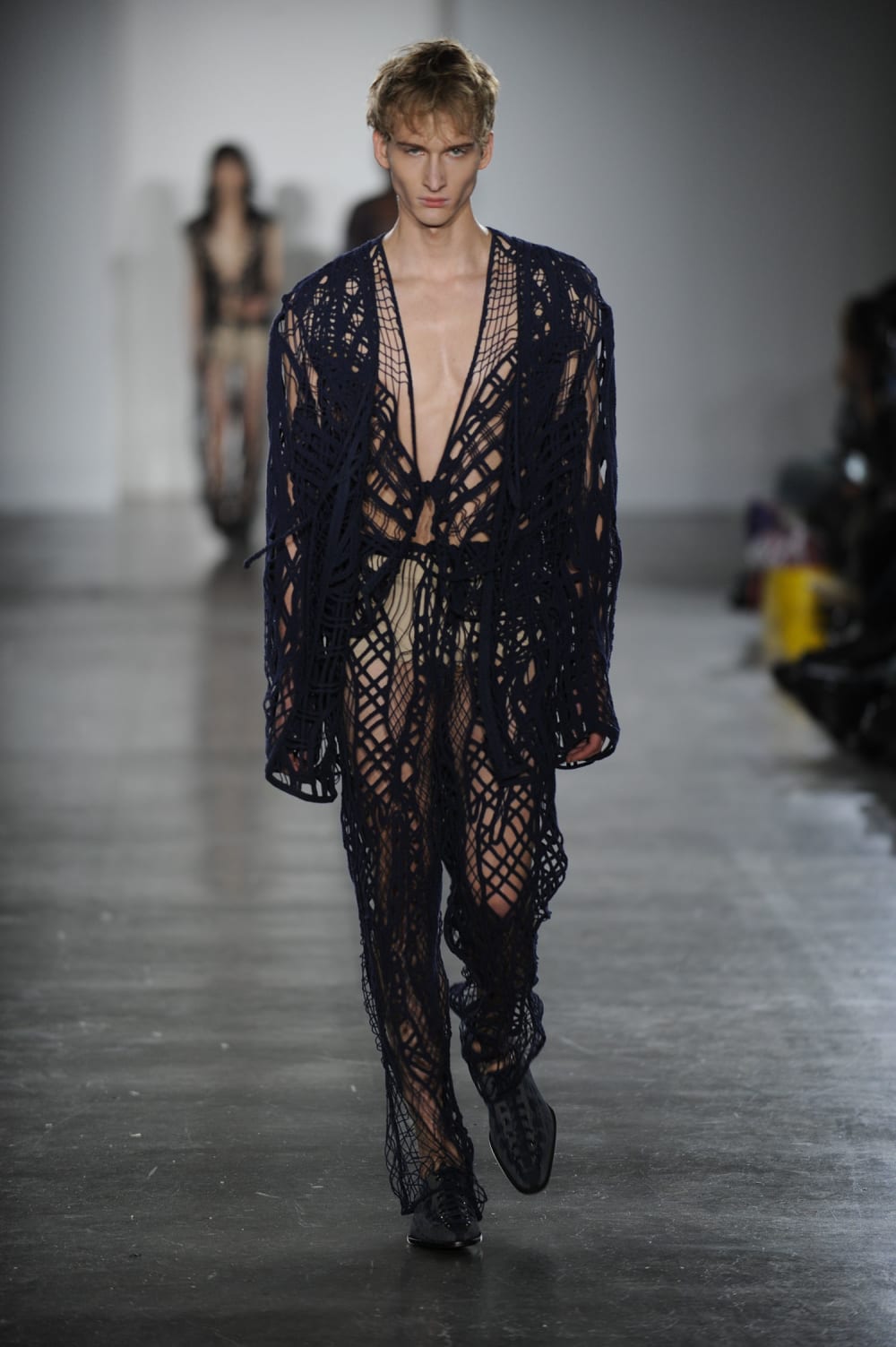 Man on runway wearing one piece navy blue crochet jumpsuit with deep v neck and black laced shoes