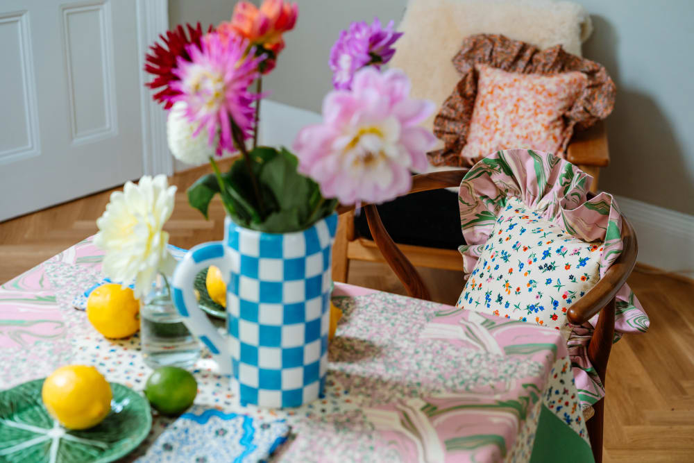 A table covered in a bright tablecloth, with a vase of flowers. In the background are 2 cushions propped on chairs