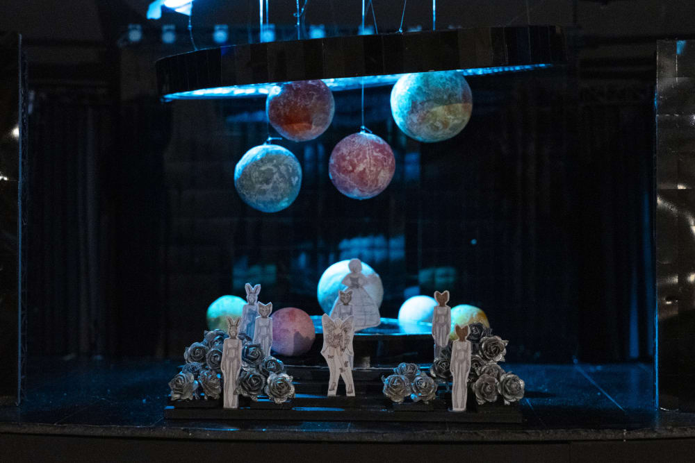 A miniature model of planets hanging from the ceiling with animal-headed humans standing underneath, surrounded by silver roses