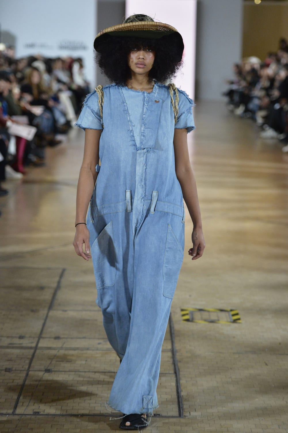 A model with a straw coloured hat on afro hair, wearing a long flowing pale blue denim dress on the catwalk