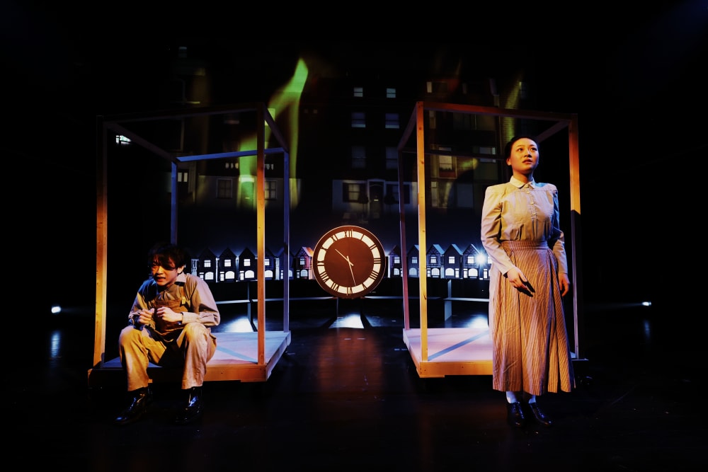 Image shows 2 people standing in front of large wooden frames – one situated on the left-hand side and one on the right-hand side. In the background is a large clock and miniature houses lit up.