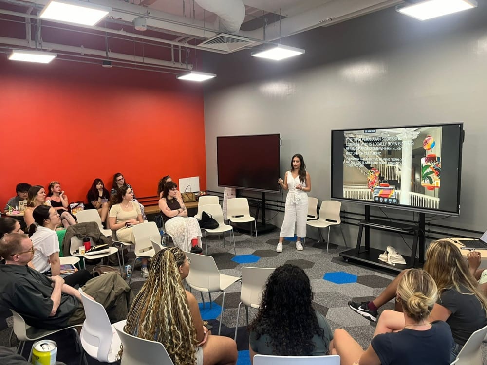 Image shows a group of people sitting in a room listening to a person who is standing presenting in front of a screen