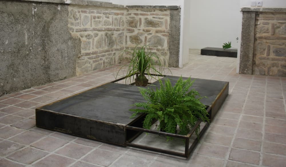 A compartmentalized metal box with two plants in, in a tiled courtyard