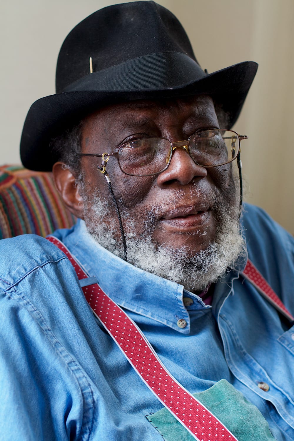 A close-up portrait photograph showing the head and shoulders of artist Frank Bowling at home wearing red braces, a blue shirt, a black hat and glasses.