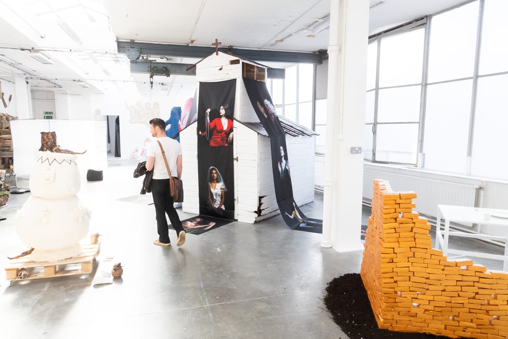 A man walks through the BA Fine Art Sculpture exhibition. To his left is a white object sitting on a wooden pallet. To the right of the shot is a low wall made of yellow bricks stacked on top of one another. He is walking past a small church-like building from which two banners hang printed with portrait photographs on a black background.