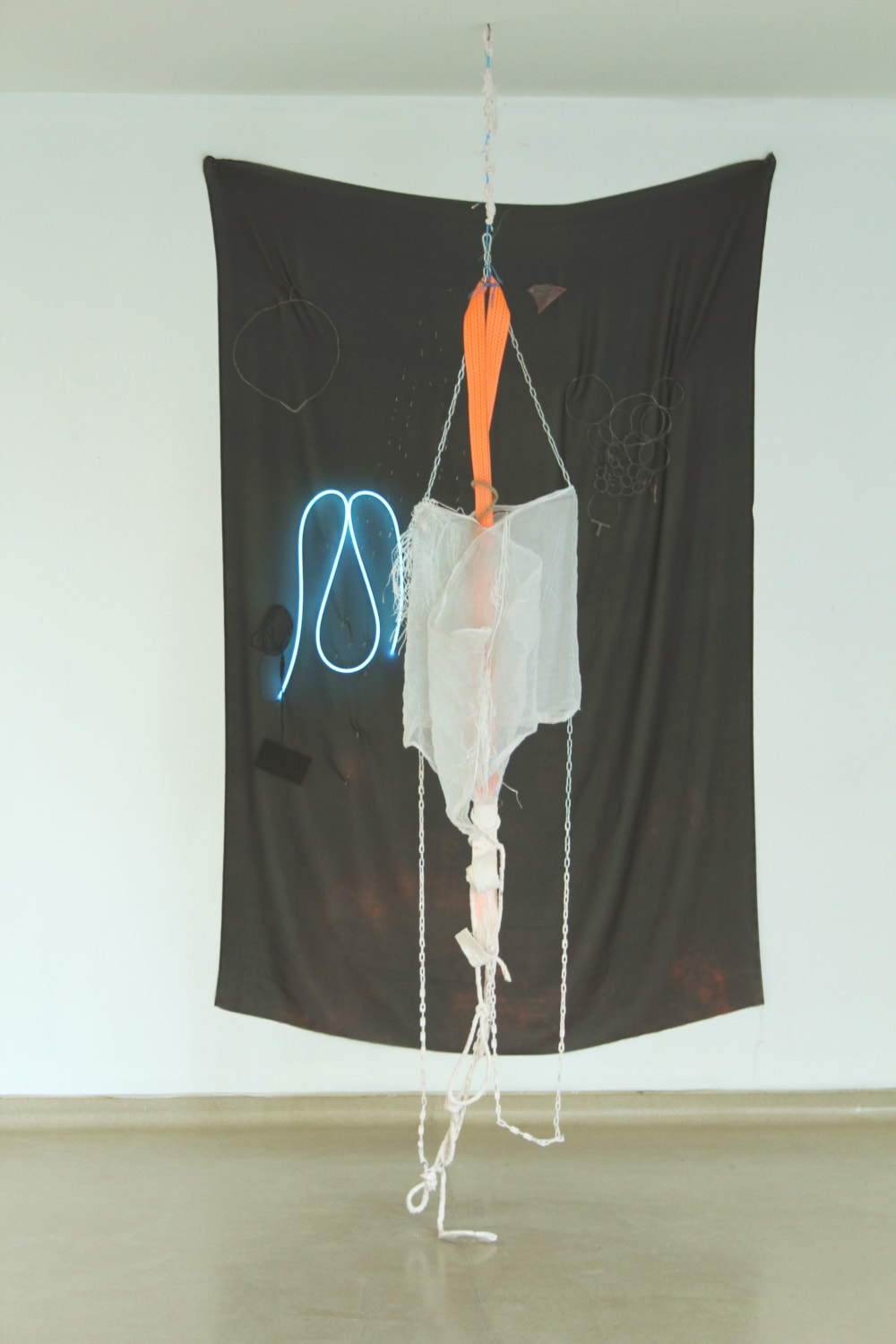 An installation image of an exhibition showing a mixed-media artwork against a while wall. The artwork is comprised of a black sheet against which some orange and white fabric, strapping and rope hangs, with a blue neon line in a curved shape to the left of the piece.