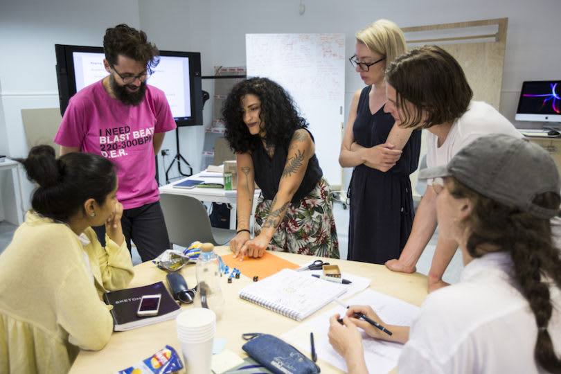 six people engaged around a work table covered in notebooks and stationery.