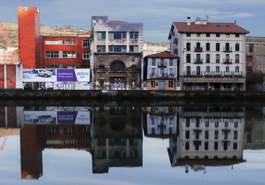 Landscape photograph of a row buildings in Bilbao. The buildings are of different heights and colours and look abandoned and old. The row of buildings are reflected in the river which is located directly in front.