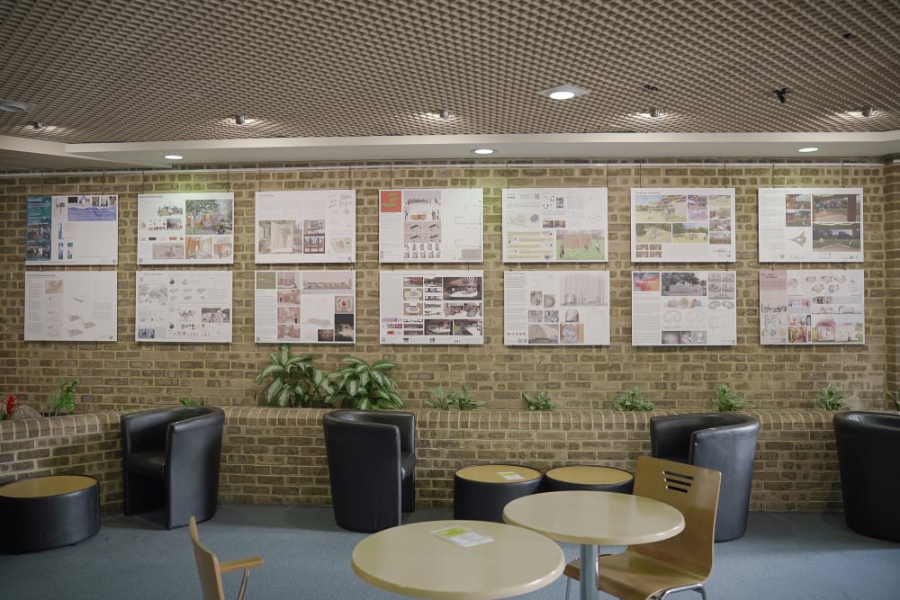 Landscape photography showing 14 A3 landscape boards each with a design proposal for the Royal Brompton Hospital hung in the hospital cafe. The boards are hung against a brick wall and in the foreground, there are tables, chairs and green plants.