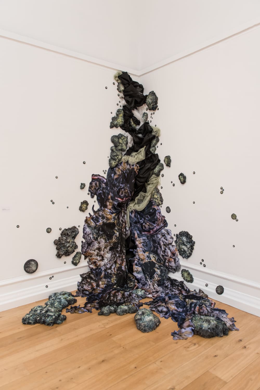 In the corner of a white room, dark fabrics and rocks look like they are infecting the room by climbing the wall and spilling out.