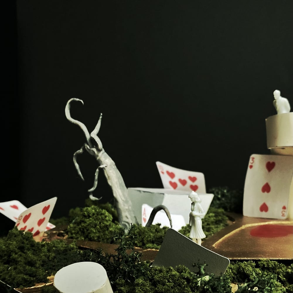 An outdoor set, with green grass and a tree branch, playing cards are positioned around the set, a female model is stood in the centre.  