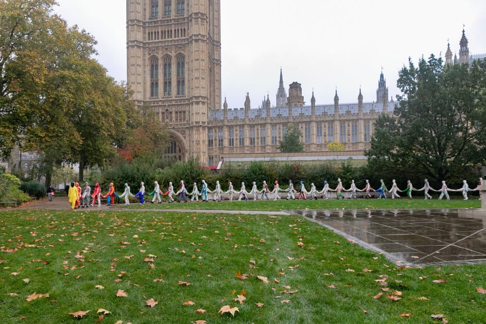 In the distance you can see a long line of people wearing connected suits walking across a grassy park. Behind them you can see a large old building, part of the Houses of Parliament.