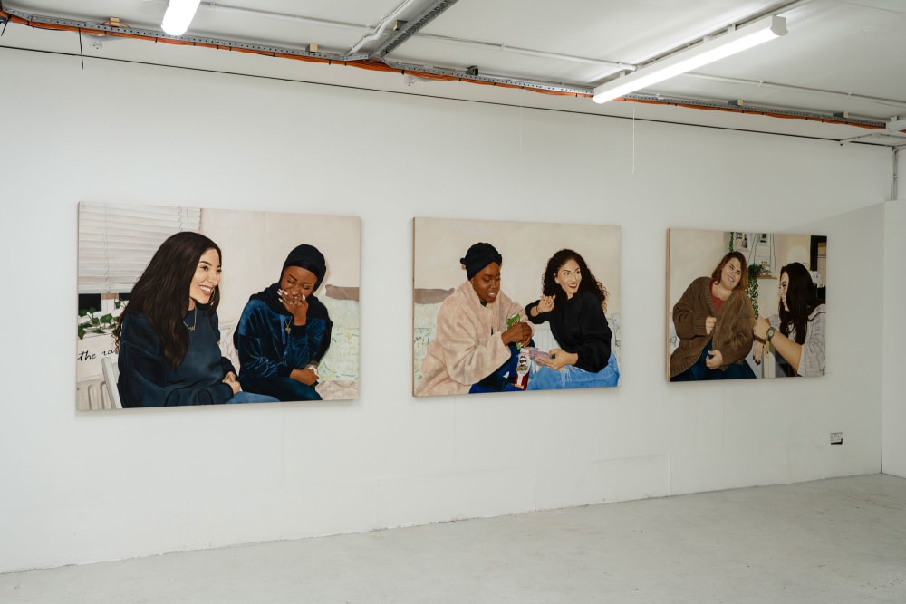 A triptych of paintings hanging on a white gallery wall showing 3 pairs of friends sitting and laughing together.