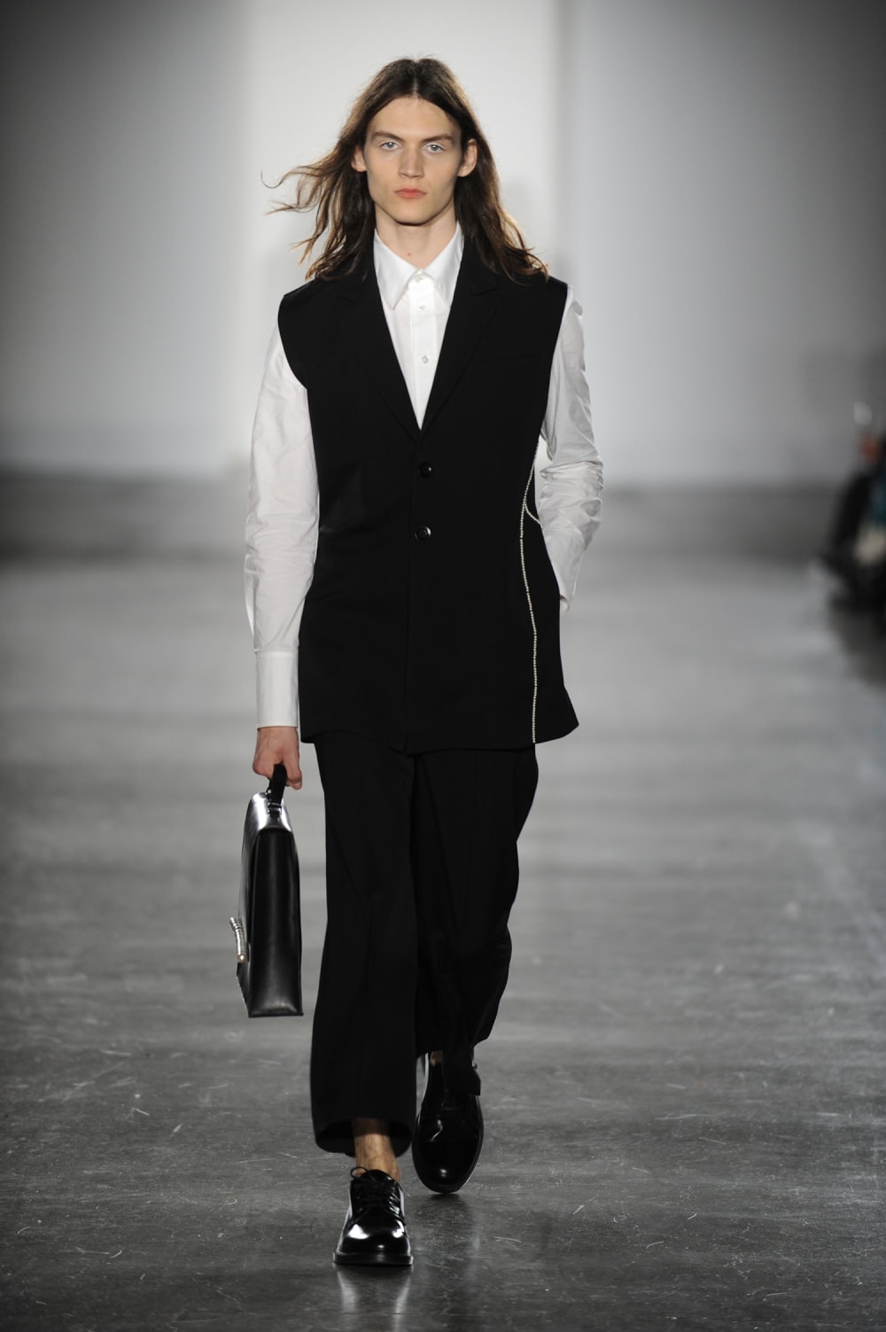 Man on runway wearing white collar smart shirt, sleeveless single breasted bottomed up jacket with white detailing on left side, black straight cut trousers, holding black briefcase, black laced smart shoes