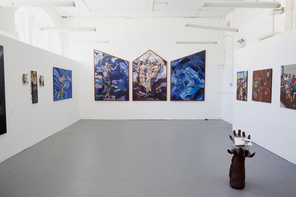 Image shows a white room with multiple paintings hanging on the wall. To the right, in the foreground, a sculpture of two open hands can be seen.