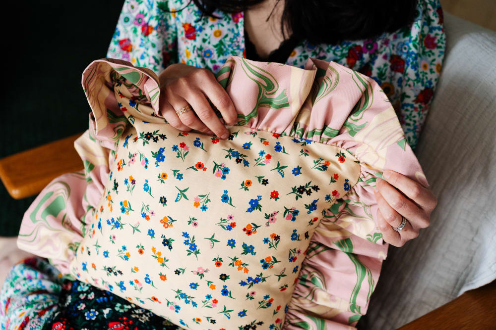 A person holding a cushion. The cushion is cream with tiny flowers in red, blue, orange and pink. The cushion has a frill around the outside which is pink with green marbling effect. 