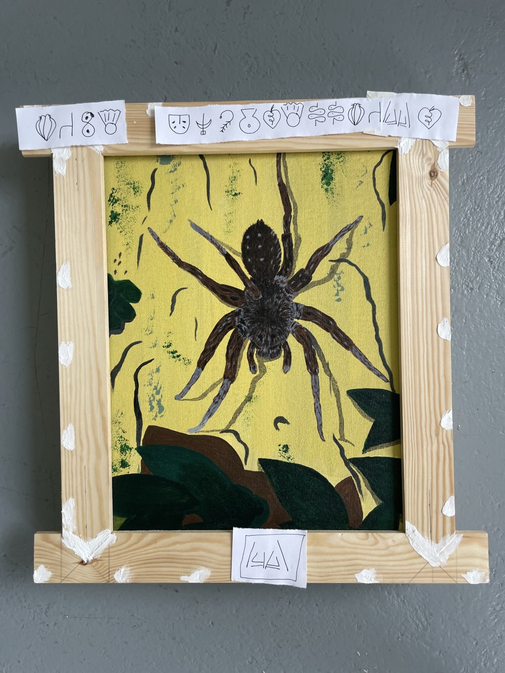Photograph of a painting of a spider with a wooden frame and symbols written on paper over the frame. 