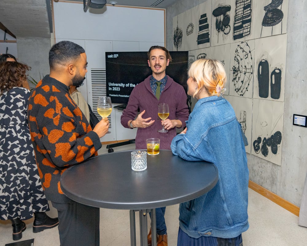 Three people stood around a table, talking. In the background there's a large, monochrome artwork on the wall 
