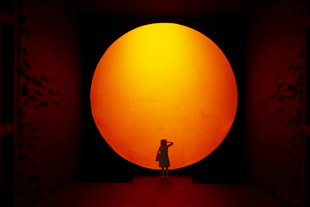 A miniature model installation of a bright sun, with a little girl in front of it covering her eyes