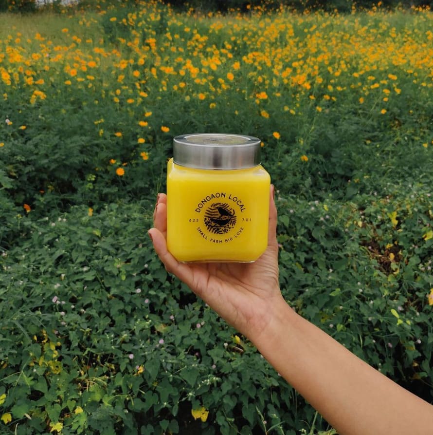 A jar of ghee being held up in front of a field with yellow flowers