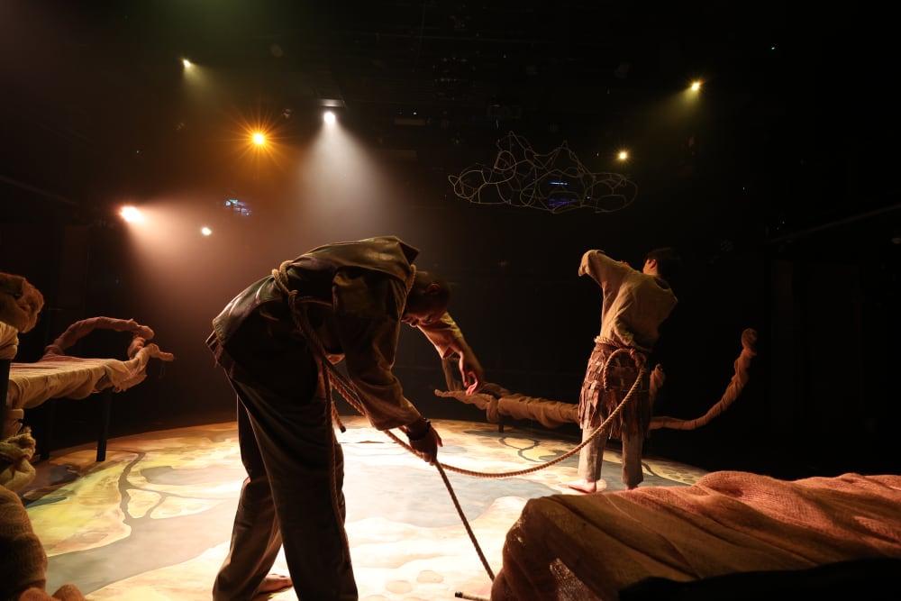 Image shows 2 people tied together with ropes moving around a stage which features different levels of benches adorned with fabric.