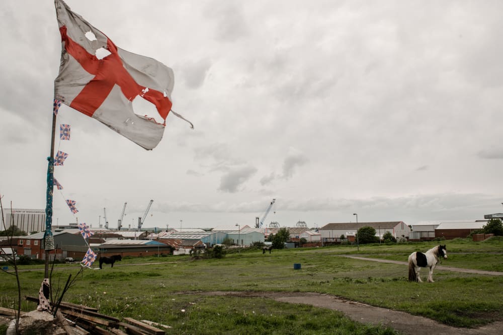In the foreground is a tattered England flag, with small union jacks tied up the pole. There's horses tethered in the field and in the background is the port of Middlesborough with buildings and cranes. Two thirds of the photo is a bright, grey sky.