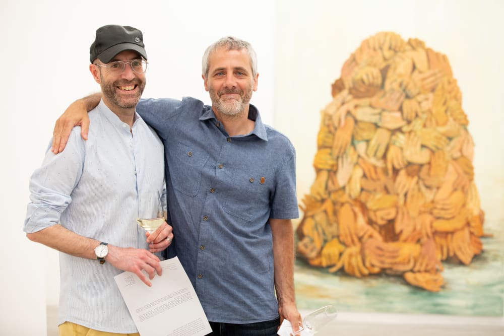 2 men with their arms around each other smiling to the camera. To the right, slightly out of focus is a painting of a pile of hands. 