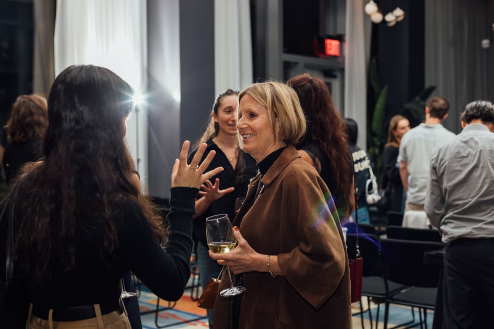 2 women chatting; one is wearing a brown suede jacket and holding a glass of wine. She's smiling at another guest, who's back is to the camera. 