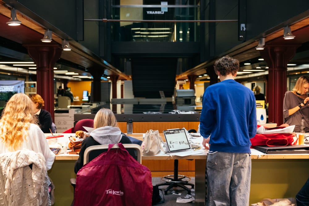 Students standing and seated at a long table in CSM library, with reading materials, bags and laptops visible.