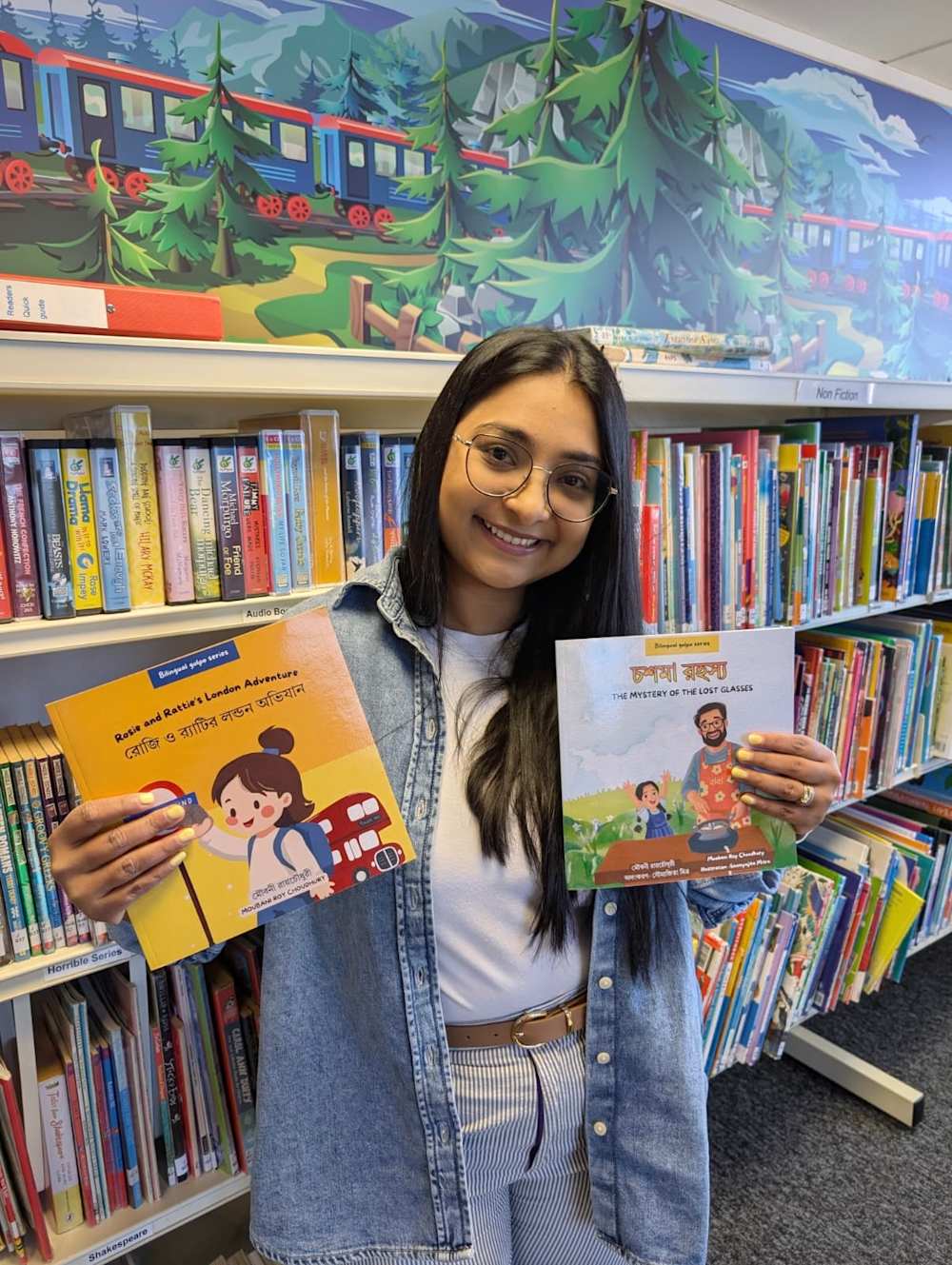 Moubani standing in a library holds two bilingual Bengali-English children’s books by Moubani Roy Choudhury in front of bookshelves filled with children’s literature.