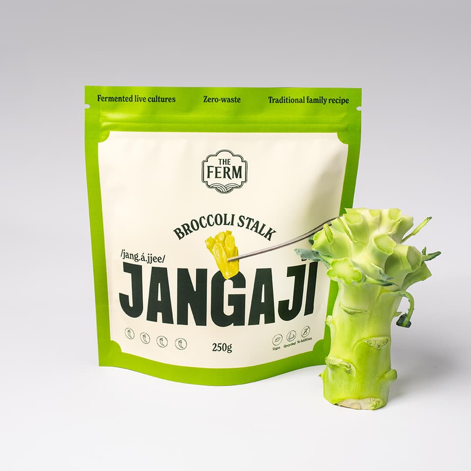 A green pouch of The Ferm's product broccoli stalk jangaji next to a green broccoli stalk.