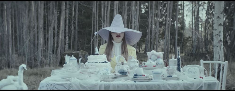 A still featuring a woman all in white, wearing in a big white hat sitting in front of a dinner table in the woods.