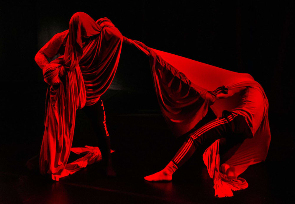 Two performers, lit in red against a black background, are wraped in draped fabric which connects them as they move through the space.