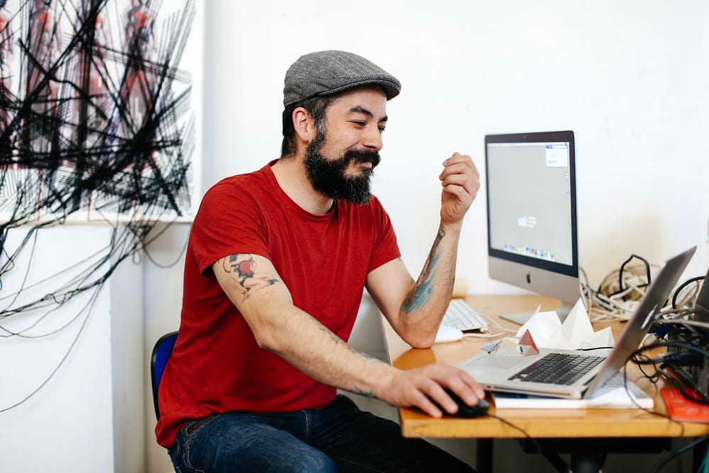 Bearded man wearing a flat cap and smiling while sitting at his laptop
