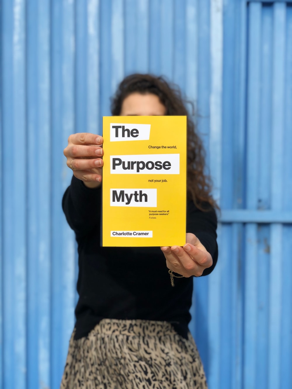 Woman holding book in front of her face. The book cover is yellow, titled 'The Purpose Myth'